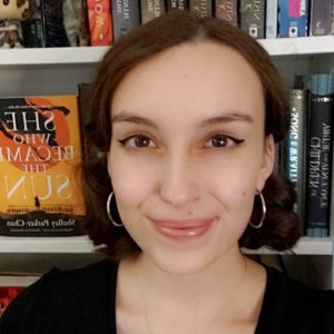 rounded photo of Ilana, a young white woman with dark brown eyes and reddish-brown hair. She has black eyeliner on and is wearing silver hoop earrings. She sits in front of a bookshelf with "She Who Became the Sun" noticeably displayed.
